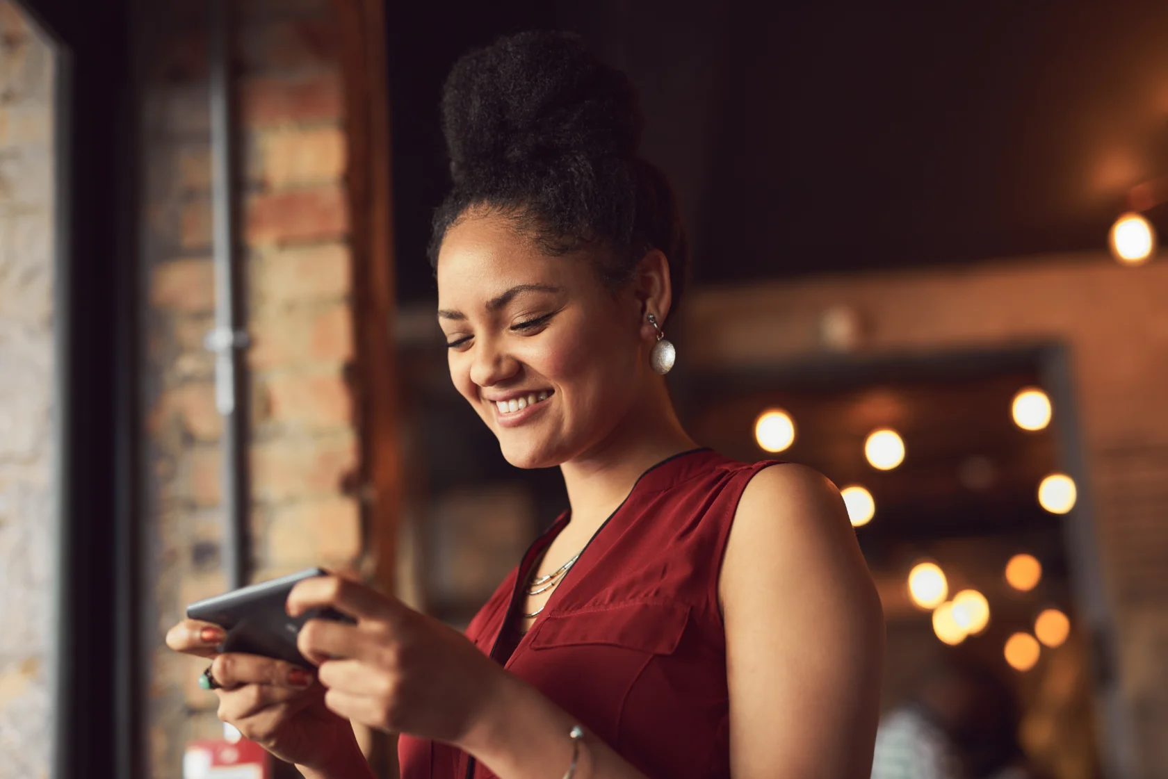 Woman in a burgundy top smiles while looking at her smartphone indoors, with warm lights and a brick wall in the background.