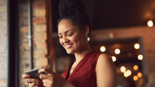 Woman in a burgundy top smiles while looking at her smartphone indoors, with warm lights and a brick wall in the background.