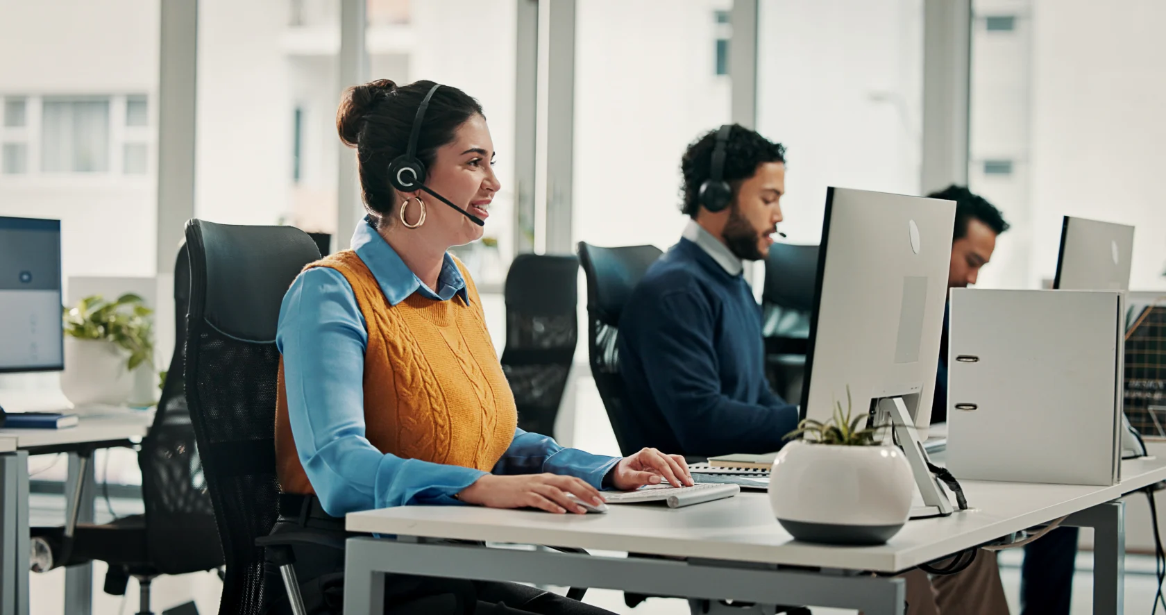 Three people wearing headsets sit at desks with computers in a modern office, working and speaking with customers.