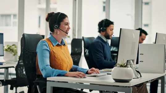 Three people wearing headsets sit at desks with computers in a modern office, working and speaking with customers.