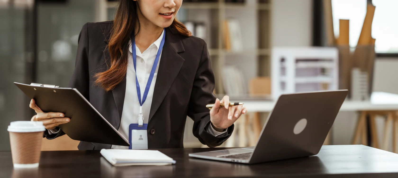 A person in business attire holds a clipboard and pen while working on a laptop at a desk with a notebook and coffee cup.