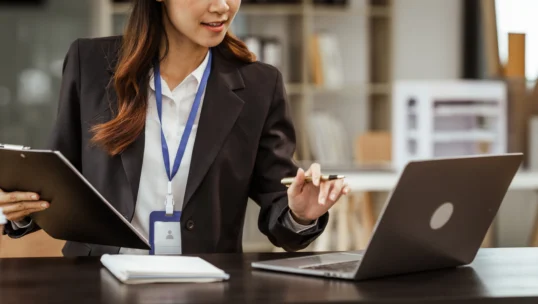 A person in business attire holds a clipboard and pen while working on a laptop at a desk with a notebook and coffee cup.