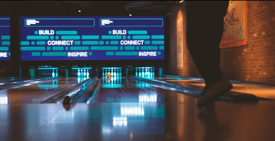 A bowling ball rolls down a lane toward pins with a digital scoreboard above displaying the words build, connect, and inspire.