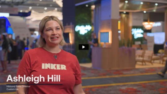 A woman in a red “INKER” shirt stands in a convention center, smiling. On-screen text reads: “Ashleigh Hill, Inbound Sales Director, Custom Ink.”.