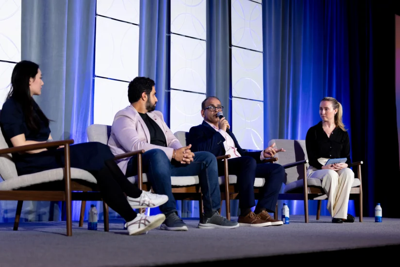 Four people sit on a panel stage having a discussion, with microphones and water bottles, in front of blue and white backdrop.