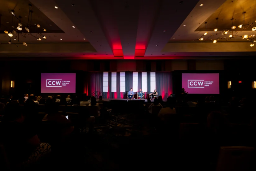 A panel discussion at a conference with three people on stage, two CCW signs on screens, and an audience seated in a dimly lit room.