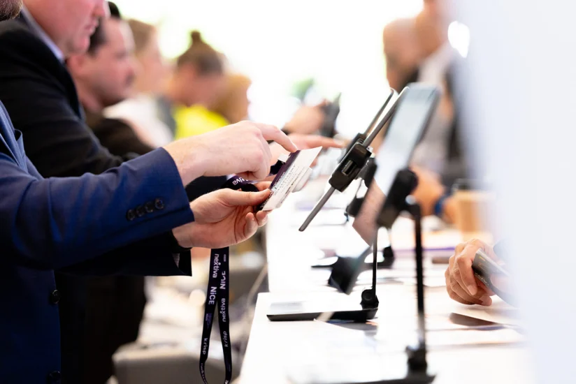 A person hands an ID badge to a staff member at a check-in counter, with tablets and lanyards visible on the desk.