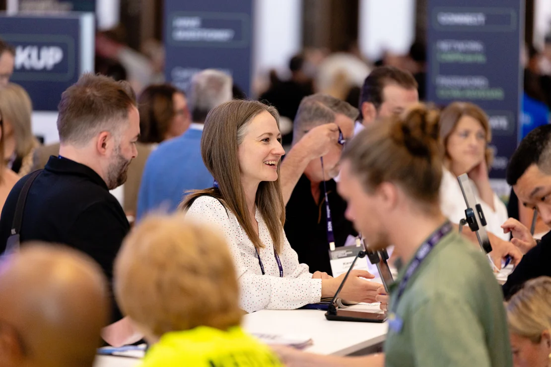 People standing and sitting at a busy conference registration desk, talking and checking in. Signs and blurred attendees are visible in the background.