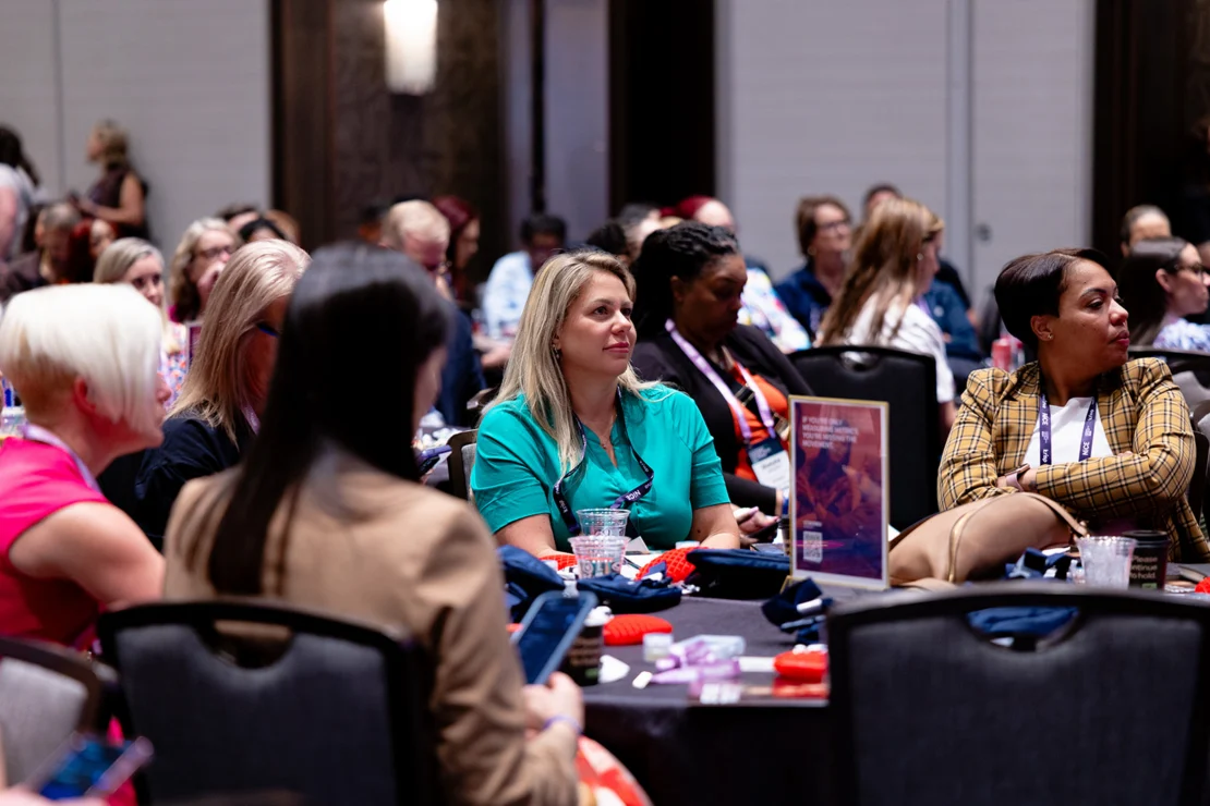 A group of women sit at tables in a conference room, listening attentively to a speaker. Conference materials and drinks are on the tables.