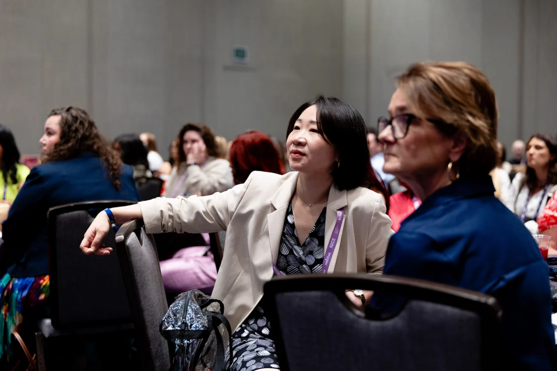 A group of people sit and listen attentively at a conference or seminar in a large room with neutral-colored walls.