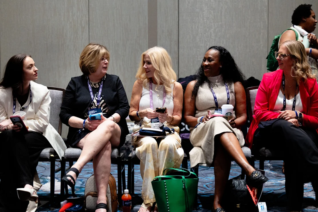 Five women sit in a row on chairs at a conference, talking and holding phones, with bags on the floor and badges around their necks.
