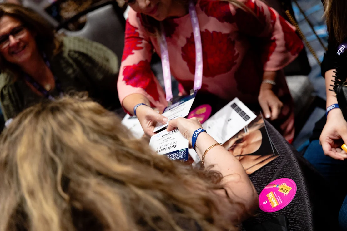 Several people sit together, one handing a name badge to another. Conference lanyards and “VIP” stickers are visible.