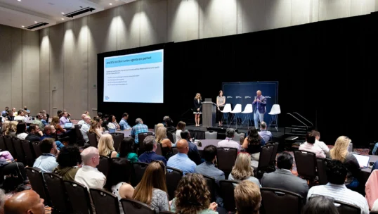 A large audience listens to three speakers onstage at a conference. A presentation slide is displayed on a screen beside them.