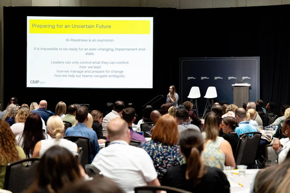 A woman stands at a podium presenting to a seated audience in a conference room, with a slide titled “Preparing for an Uncertain Future” displayed on a large screen behind her.