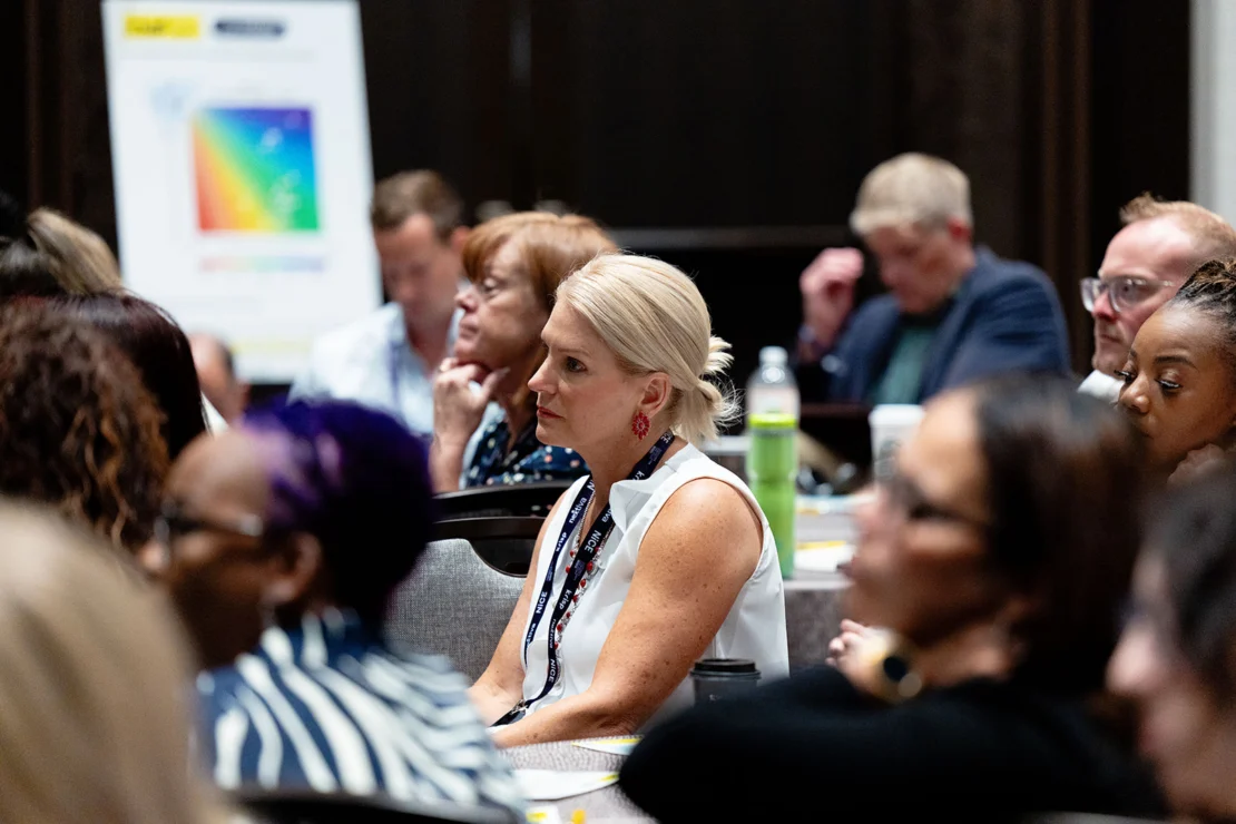 A group of people sit and listen attentively during a conference or seminar; a colorful chart is visible on a board in the background.