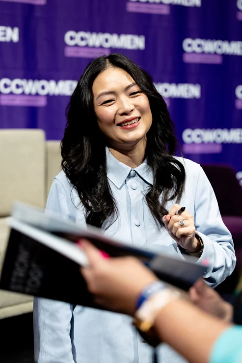 Woman with long black hair, wearing a light blue shirt, smiles and holds a pen while interacting with someone in front of a purple “CCWOMEN” event backdrop.