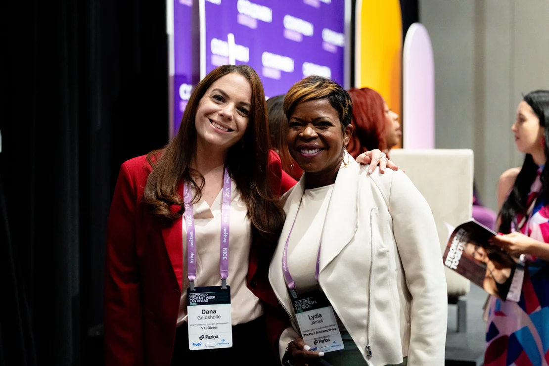 Two women wearing conference badges smile and pose together at an event, with other attendees and a purple event banner visible in the background.
