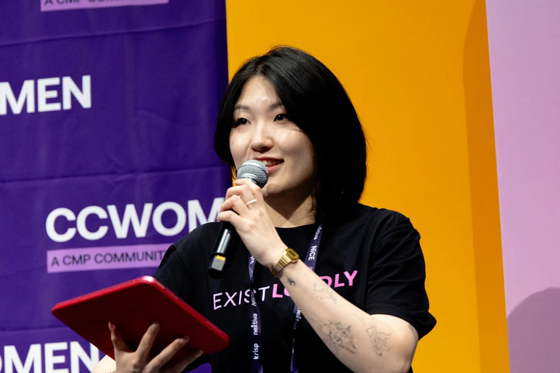 A woman holding a microphone and red tablet speaks on stage at an event, with CCWOMEN signage and a bright yellow-purple background behind her.