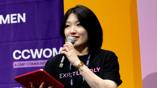 A woman holding a microphone and red tablet speaks on stage at an event, with CCWOMEN signage and a bright yellow-purple background behind her.