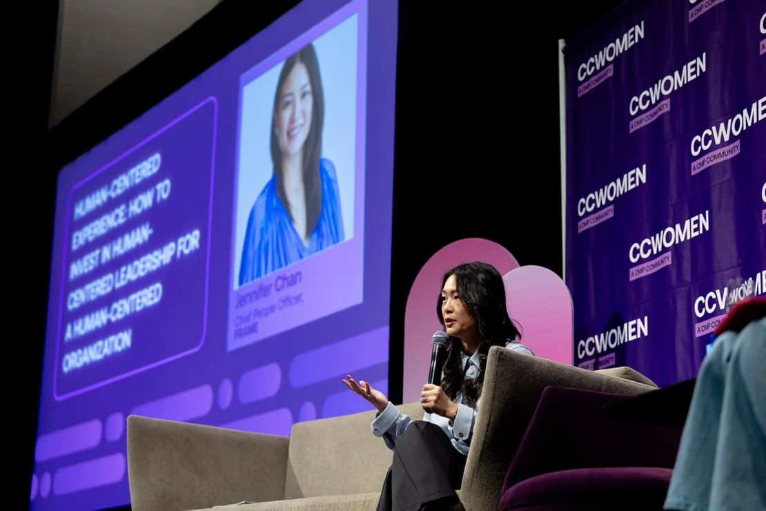 A woman speaks into a microphone while seated on stage; behind her is a projector screen with her photo and presentation details, and a purple CCWOMEN backdrop.