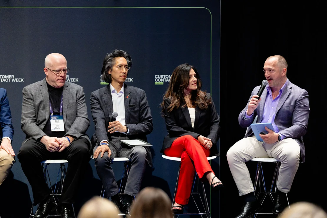 Four people sit on stools participating in a panel discussion at Customer Contact Week; one person speaks into a microphone while holding a tablet.