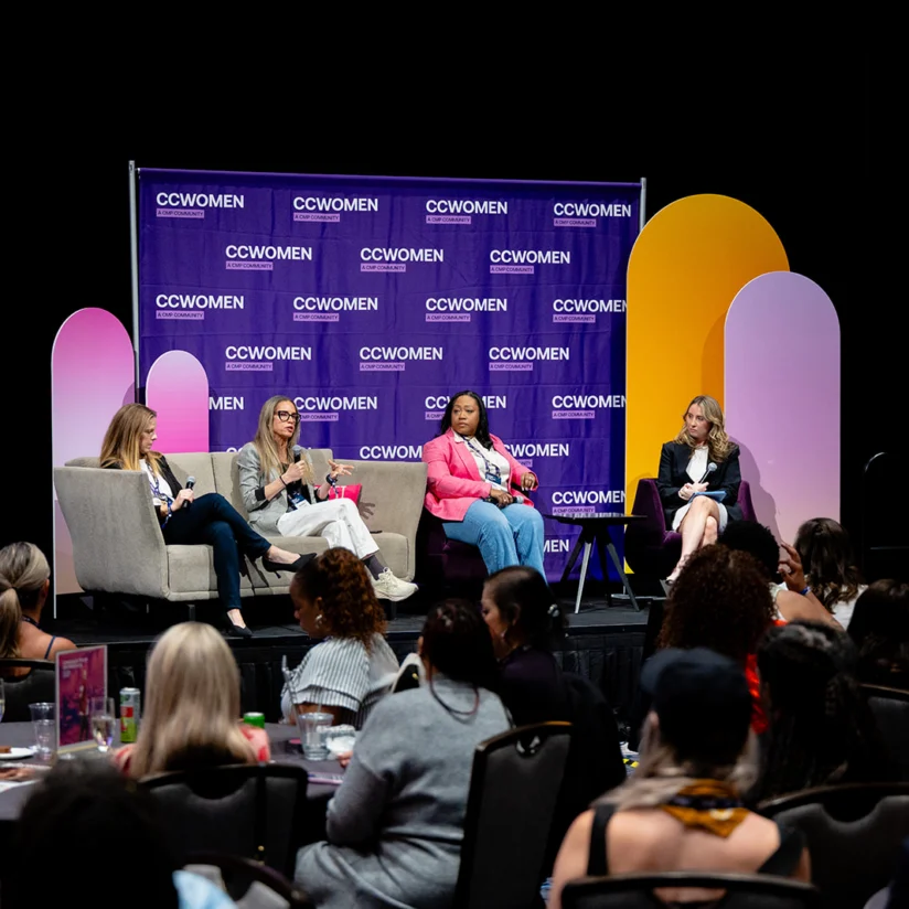 Four women sit on a stage having a panel discussion at a CCWomen event, with an audience seated at tables in the foreground.