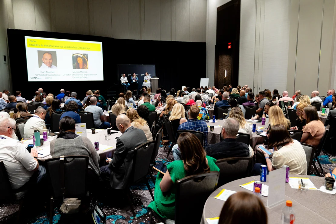 A large group of people attends a conference in a hotel ballroom, watching a panel discussion on a stage beneath a screen displaying speaker information.