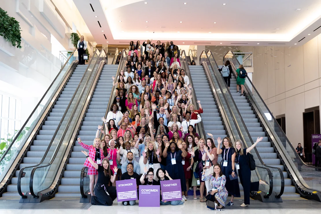 A large, diverse group of women pose at the base of escalators in a convention center, holding up purple signs that read “Stronger Together” and “Crowd Women,” smiling at the camera.