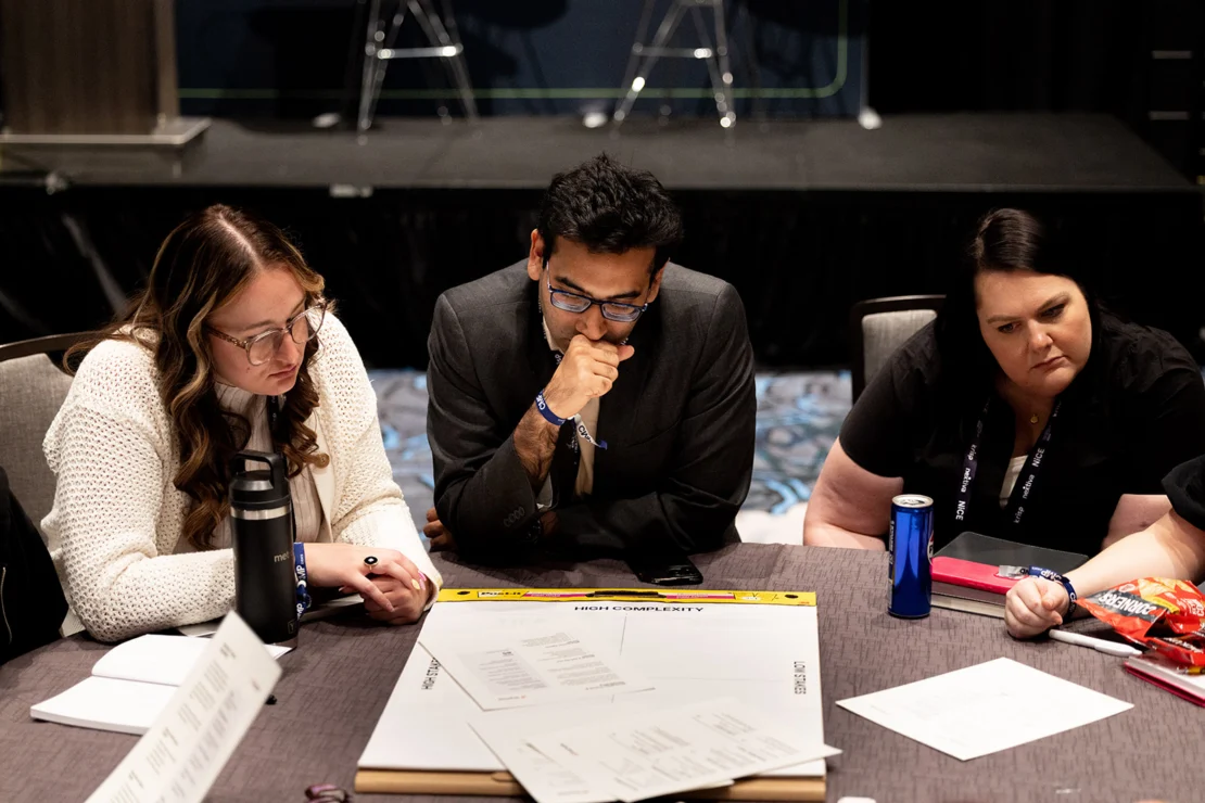 Three people sit at a table looking at documents, appearing focused and engaged in discussion. Snacks and drinks are visible on the table.