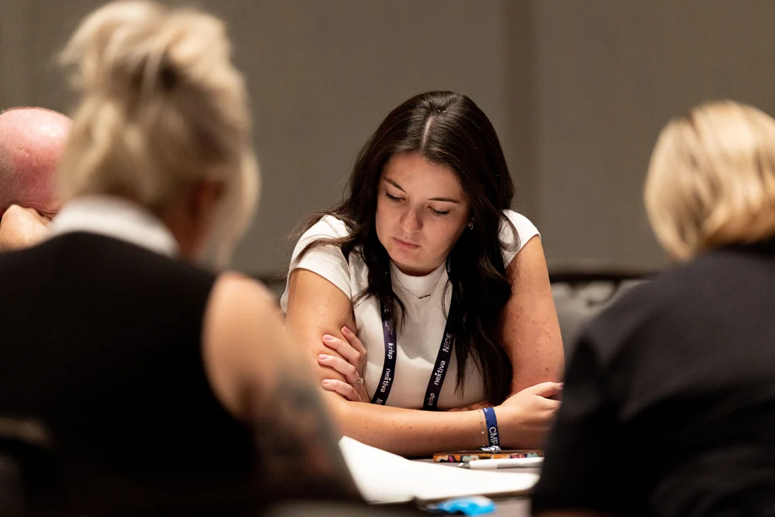 Woman with long dark hair sits at a table, looking down and reading papers, surrounded by other people in a meeting or workshop setting.
