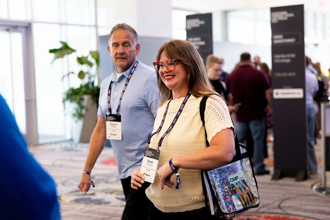 Two people wearing conference badges walk indoors at an event. The woman carries a colorful tote bag, and people are visible in the background near informational signs.