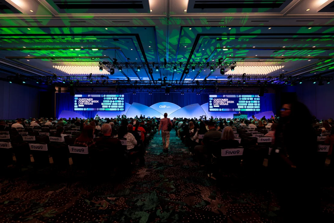 A large audience sits in a conference hall facing a stage with screens displaying “Customer Contact Week” under green and blue lighting.