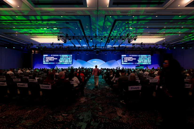 A large audience sits in a conference hall facing a stage with screens displaying “Customer Contact Week” under green and blue lighting.