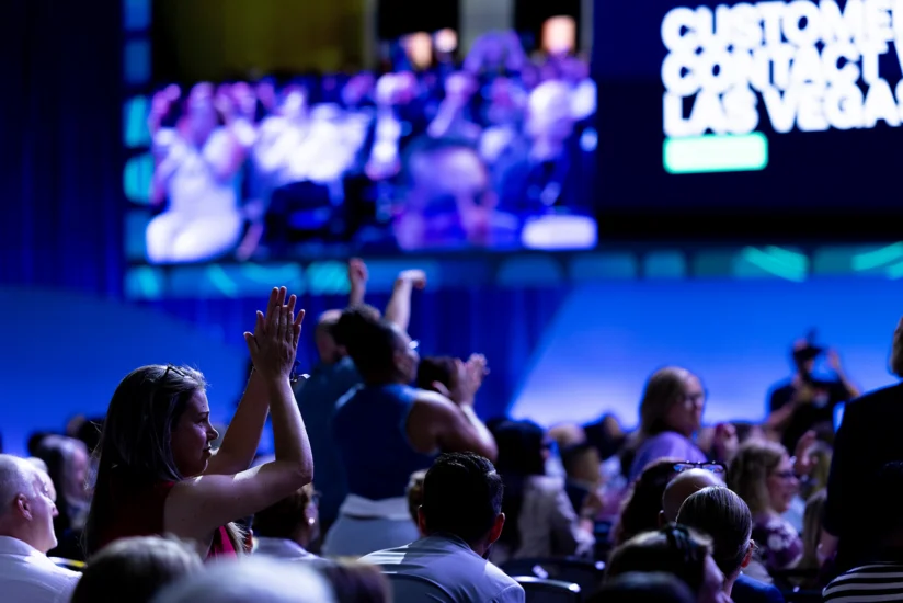Audience members, some standing and clapping, attend an event in a large room with a big screen displaying text in the background.