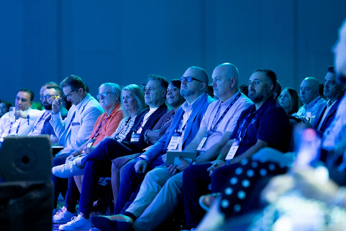 A group of people sits in rows at an indoor event, attentively facing the stage; most are wearing business attire and name badges.