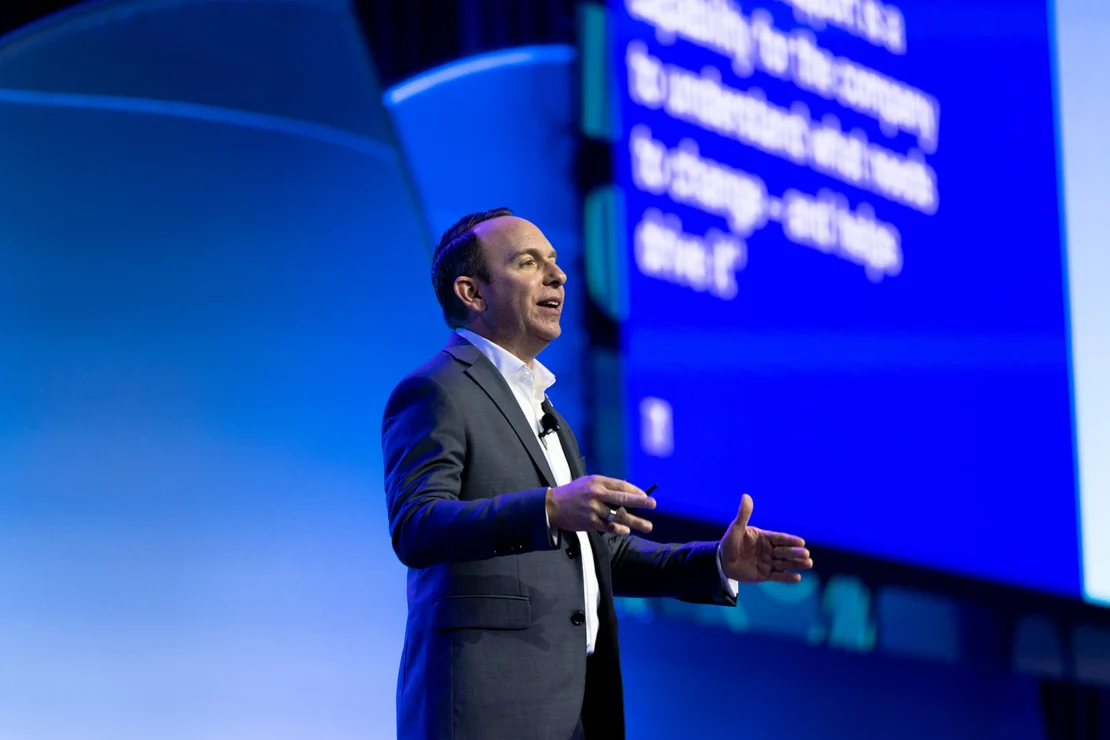 A man in a suit gestures while speaking on stage with a blue background and large screen displaying white text behind him.