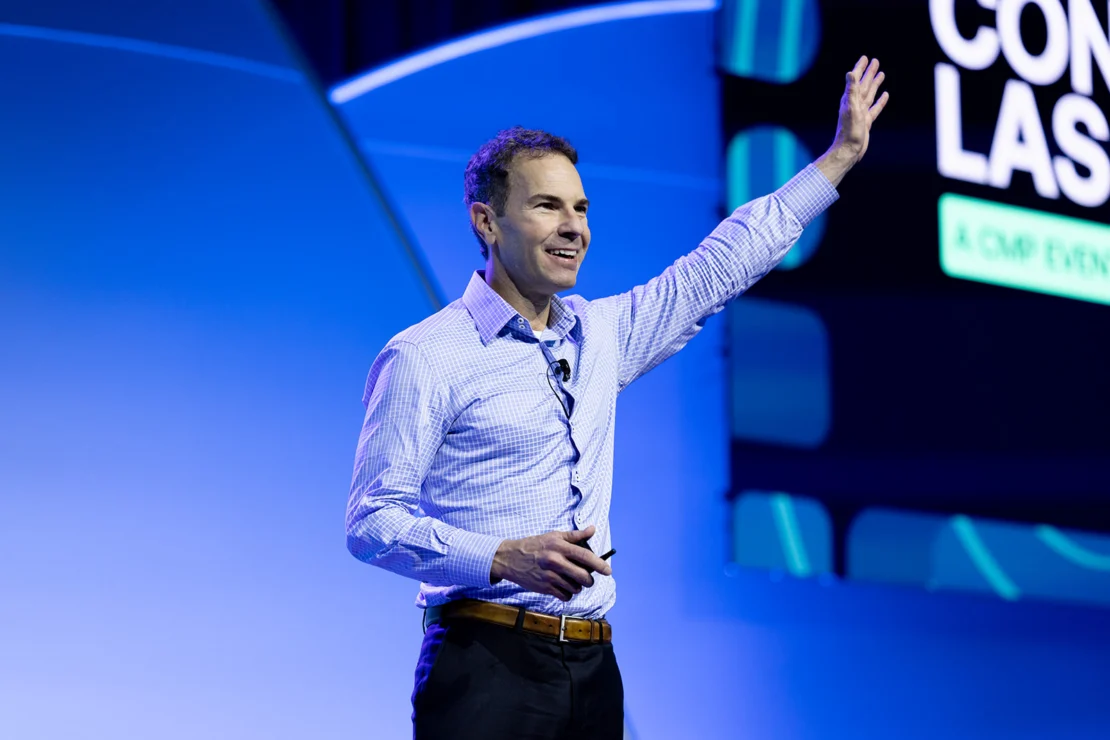 A man in a dress shirt stands on stage, smiling and raising one arm while speaking at a conference with a blue background.