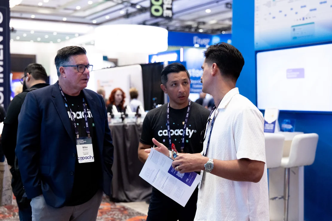 Three men have a discussion at a conference booth; two wear “capacity” shirts and badges, one holds papers, with event signage and screens visible in the background.
