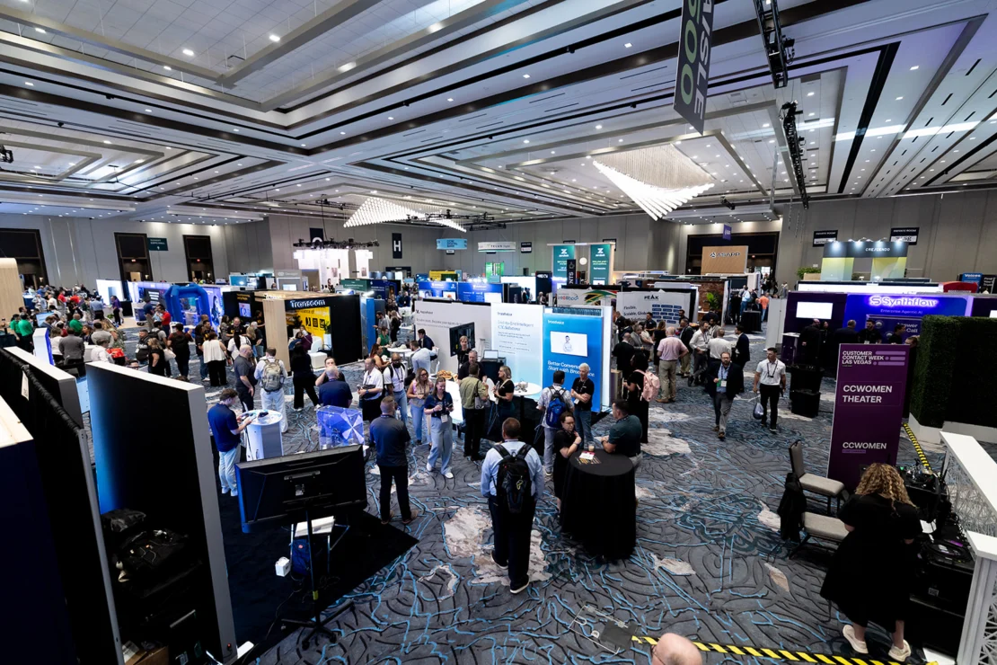 Large indoor convention with numerous booths, digital displays, and many people walking, talking, and networking in a spacious, well-lit exhibition hall.
