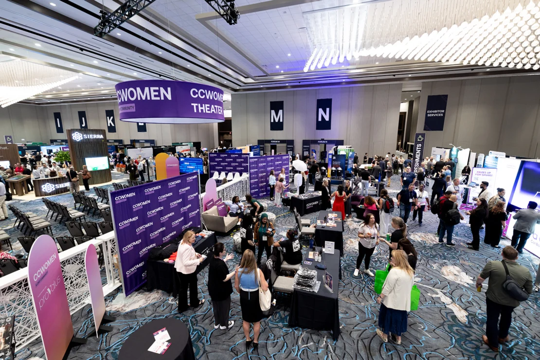 Large conference hall with various booths, banners reading “CCWOMEN THEATER,” and attendees walking, conversing, and visiting displays under bright lighting.
