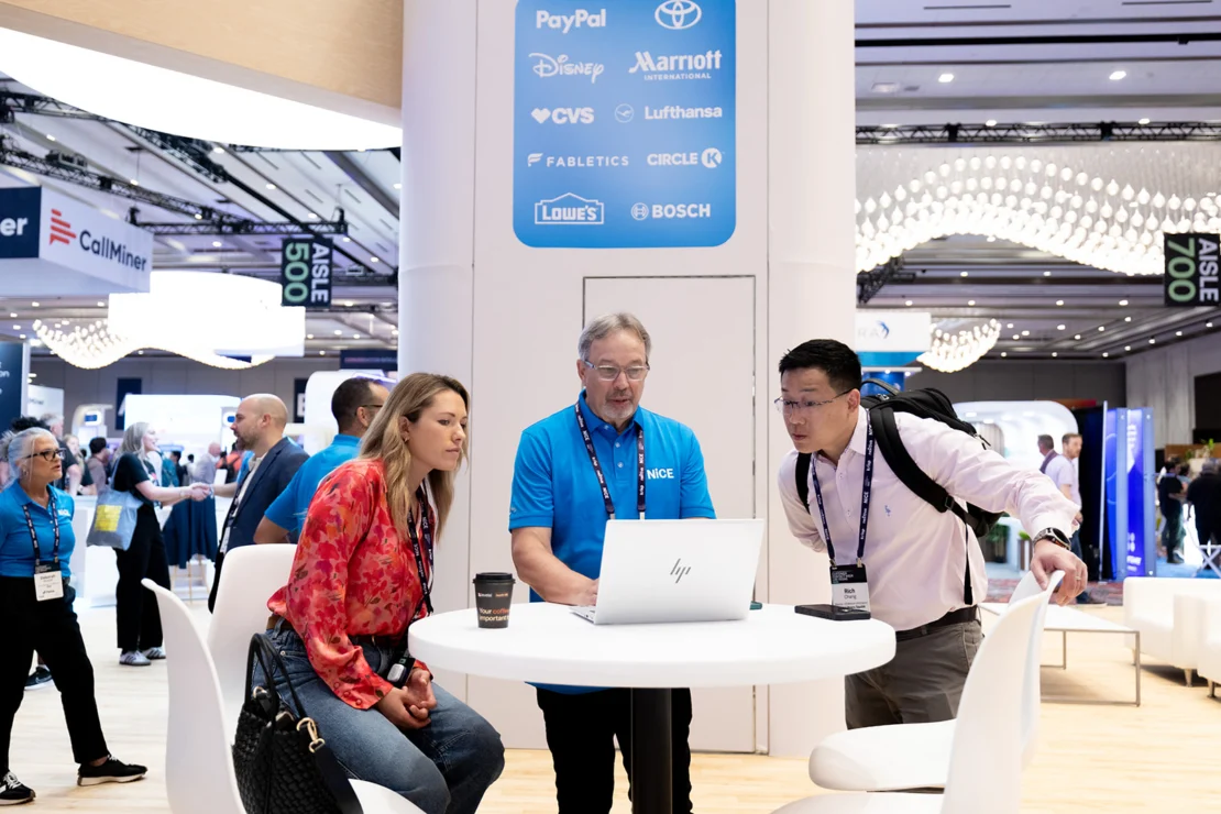 Three people gather around a round table, looking at a laptop in a busy conference hall with sponsor logos displayed on a pillar behind them.