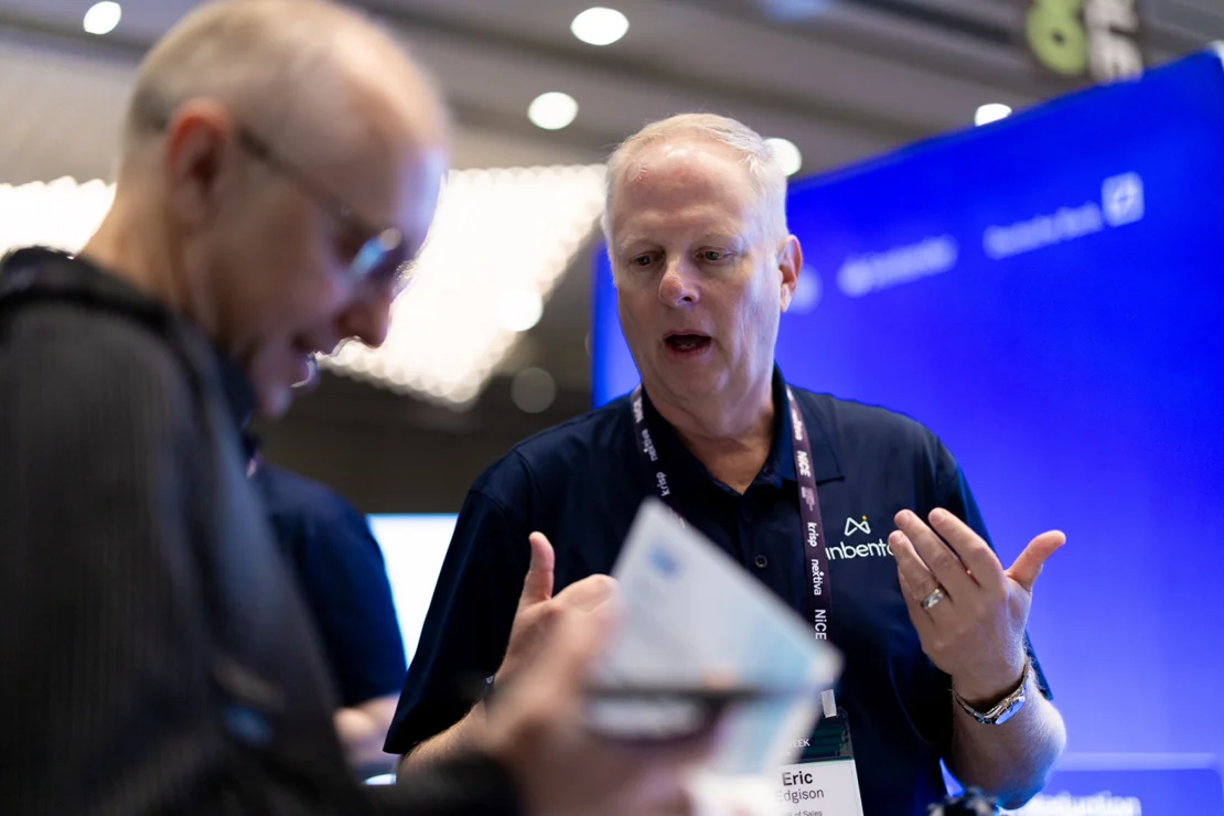 Two men interact at a conference booth, one holding papers and the other gesturing, with name badges and a blue display in the background.