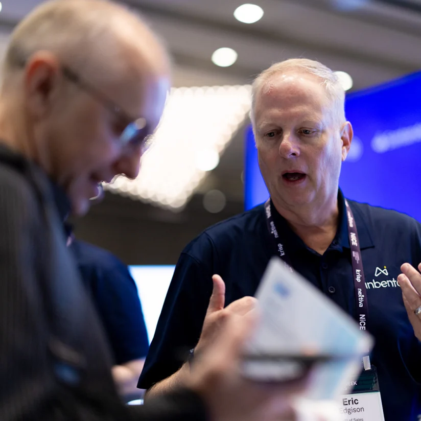 Two men interact at a conference booth, one holding papers and the other gesturing, with name badges and a blue display in the background.