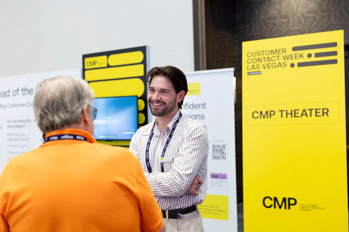 Two men talk at a Customer Contact Week Las Vegas event near a yellow sign for CMP Theater. One wears an orange shirt, the other a striped shirt and conference badge.
