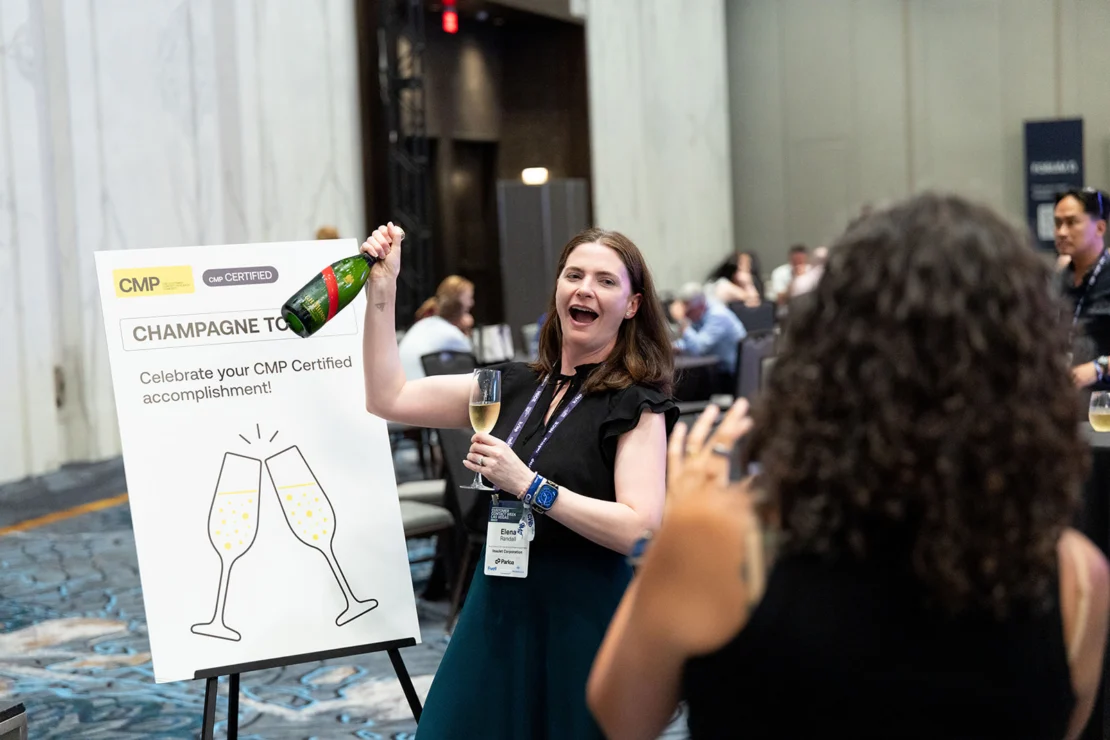 A woman opens a champagne bottle and smiles at an event beside a sign that reads “Champagne Toast. Celebrate your CMP Certified accomplishment!” Other people are present in the background.