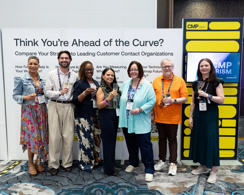 Seven people stand in front of a conference display about customer contact organizations, holding drinks and smiling for a group photo.