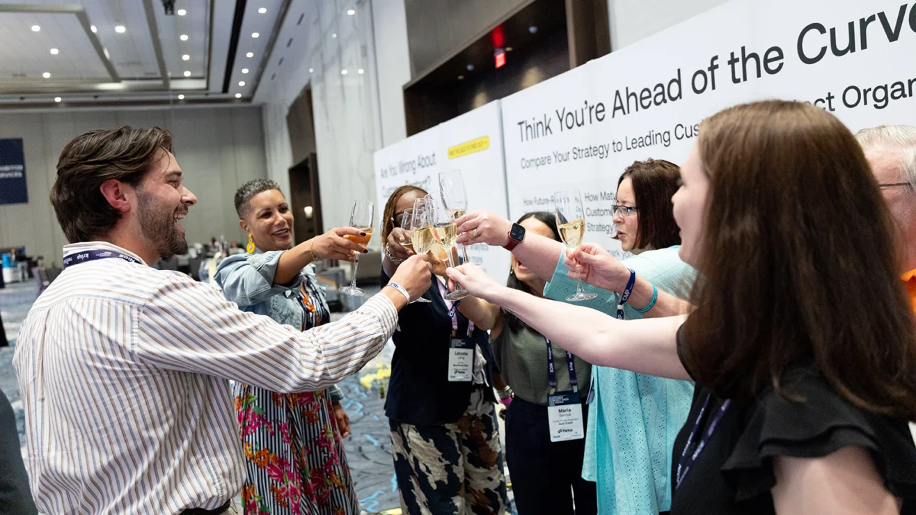 A group of people stands in a circle and toasts with drinks at a conference event, with a sign in the background reading “Think You’re Ahead of the Curve.”.