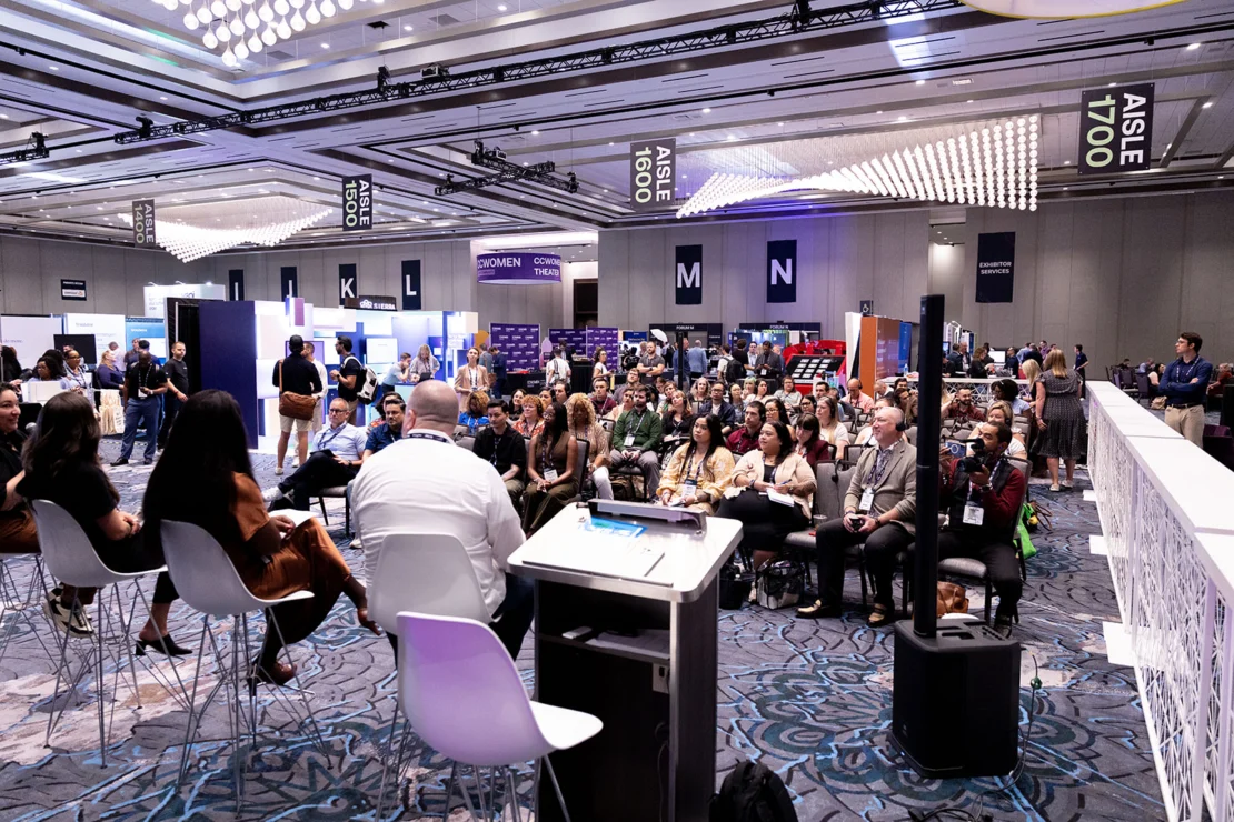 A panel of speakers addresses a seated audience at a conference in a large, well-lit room with various aisle signs and booths visible.