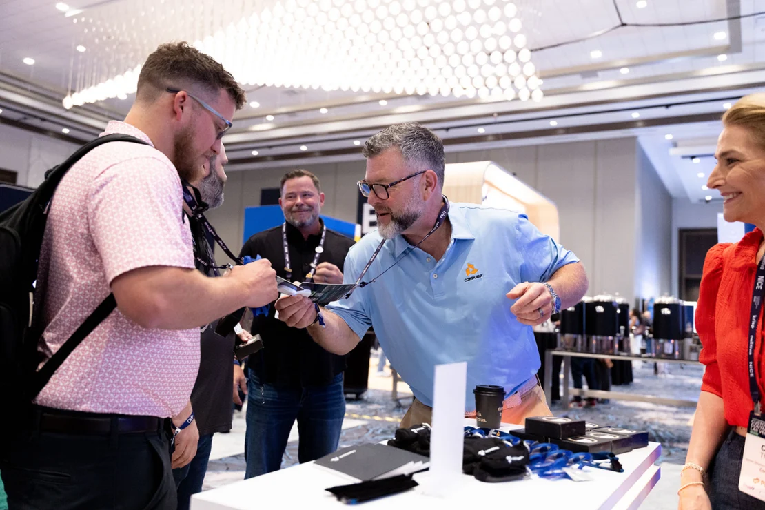 A group of people at a conference booth interact while one man hands a badge lanyard to another.