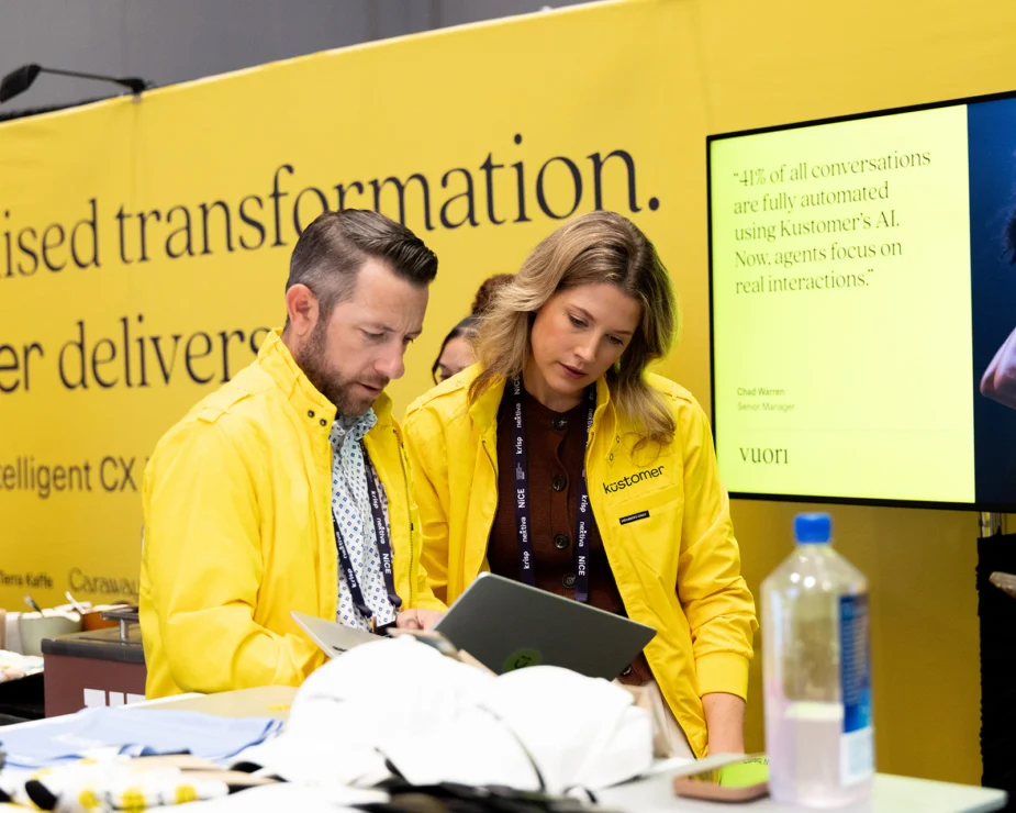 Two people in yellow jackets stand at a table, looking at a laptop. Behind them is a yellow wall with text and a screen displaying a quote about AI and customer interactions.